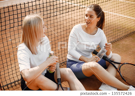 Two young women sitting by tennis net. Having conversation after game.Summer, sports, leisure. 135572426