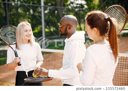 Girl friends are having tennis lesson. Black man coach talking and explaining how to play. Outdoor playground. 135572460