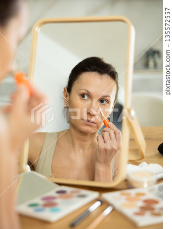 Woman applying concealer to under-eye area in front of mirror Woman applying concealer to under-eye area in front of mirror 135572557