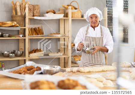 Aged female baker mixing ingredients for dough in bowl 135572565