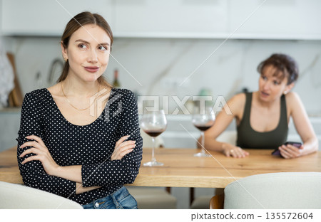 Upset middle-aged woman sitting at the kitchen table with her back to another lady quarreling to her 135572604