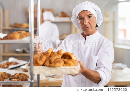 Female baker placing tray of fresh croissants on rack Female baker placing tray of fresh croissants on rack 135572660