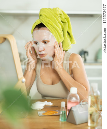 Middle-aged woman applying tissue mask on her facial skin sitting in front of the mirror Middle-aged woman applying tissue mask on her facial skin sitting in front of the mirror 135572677