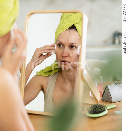 Woman with towel on head cleansing face with cotton pads Woman with towel on head cleansing face with cotton pads 135572728