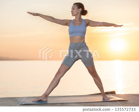 Active experienced woman teacher performs yoga asana on mat on seashore at sunset. Concept of healthy lifestyle 135572733