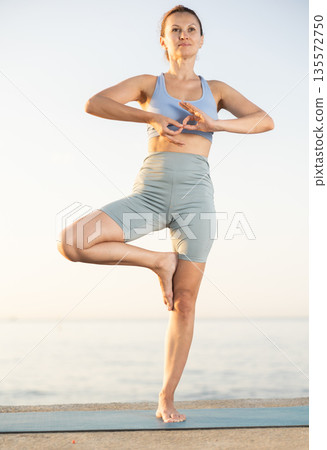 Happy woman in sportswear exercising on beach at morning, standing in warrior one leg pose on fitness mat 135572750