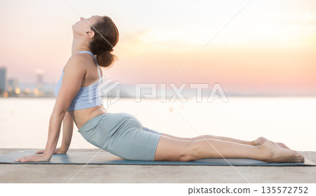 Sportive woman performing cobra pose of yoga during training on beach at sunrise 135572752