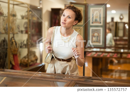 Portrait of a confident woman with an information booklet, standing in the hall of the historical museum next to the exhibit, located in glass cabinet Portrait of a confident woman with an information booklet, standing in the hall of the historical museum next to the exhibit, located in glass cabinet 135572757