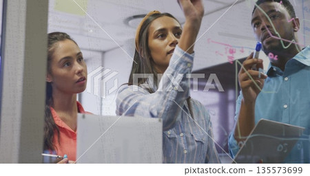 Brainstorming three professionals collaborating on glass board in office using markers and tablet 135573699