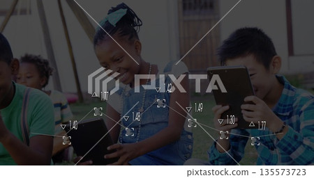 Smiling girl in denim overalls and teal bow tapping tablet on grassy lawn, with friends Smiling girl in denim overalls and teal bow tapping tablet on grassy lawn, with friends 135573723