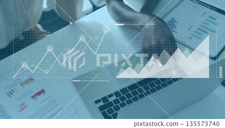 Two business women reviewing data at office desk with laptop, report, tablet and graph overlays 135573740