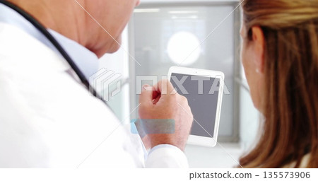 Guiding doctor wearing coat and stethoscope showing senior patient tablet screen in clinic corridor Guiding doctor wearing coat and stethoscope showing senior patient tablet screen in clinic corridor 135573906