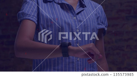 Woman in blue and white pinstripe shirt checking time on black smartwatch against brick wall 135573985