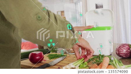 Home cook in green jacket lifting vegetable scraps into green-rim compost bin on kitchen counter 135574090