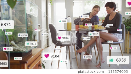 Two friends pouring juice, holding pastries in home dining area by glass doors, with media icons Two friends pouring juice, holding pastries in home dining area by glass doors, with media icons 135574093