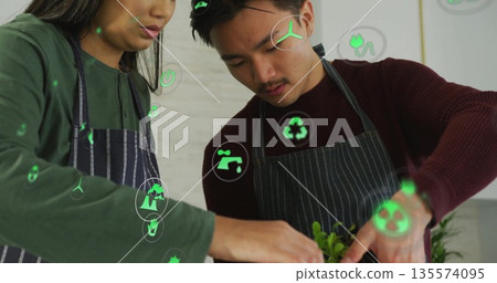 Arranging green seedlings in planter by Asian team in aprons at studio countertop with eco icons Arranging green seedlings in planter by Asian team in aprons at studio countertop with eco icons 135574095