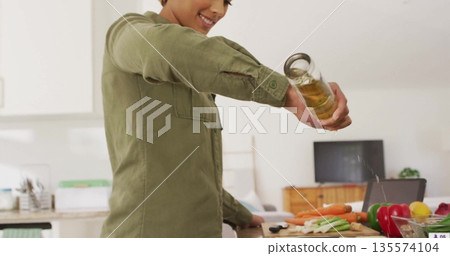 Woman pouring oil from jar onto vegetables on cutting board in home kitchen, with knife, tablet Woman pouring oil from jar onto vegetables on cutting board in home kitchen, with knife, tablet 135574104