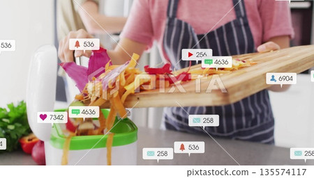 Home cook in striped apron pushing peelings into kitchen juicer at home, with social media icons Home cook in striped apron pushing peelings into kitchen juicer at home, with social media icons 135574117