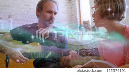 Strumming father teaching daughter guitar chords on living room sofa, with notebook and eyeglasses Strumming father teaching daughter guitar chords on living room sofa, with notebook and eyeglasses 135574191