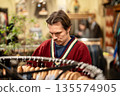 Thoughtful man customer in red sweater choosing shirts on hanger in second hand apparel store. 135574905