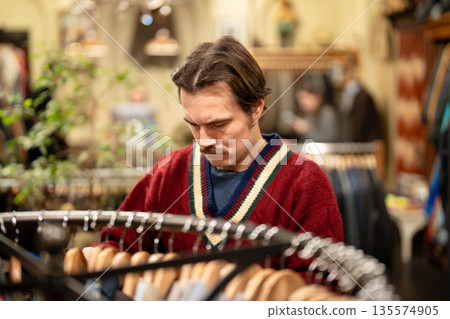 Thoughtful man customer in red sweater choosing shirts on hanger in second hand apparel store. Thoughtful man customer in red sweater choosing shirts on hanger in second hand apparel store. 135574905