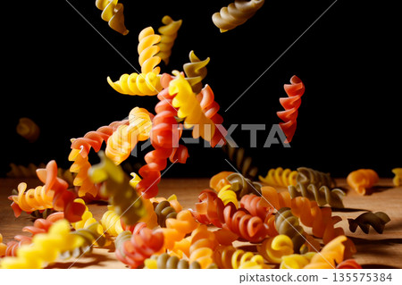 Raw tricolor fusilli pasta frozen in motion, falling onto a wooden surface with a black background Raw tricolor fusilli pasta frozen in motion, falling onto a wooden surface with a black background 135575384