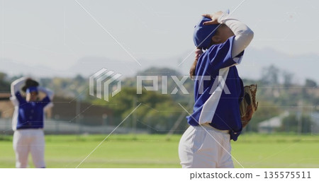 Adjusting baseball player wearing cap holding glove on field with chainlink fence, copy space 135575511