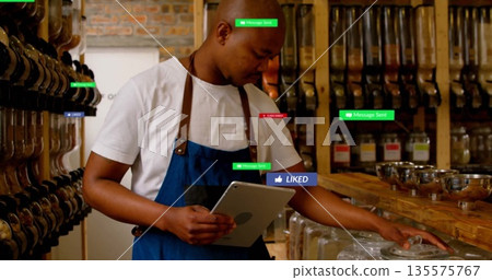 Holding tablet man wearing apron opening glass jar at bulk-food counter, with stainless steel bowls 135575767