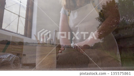 Pressing and folding dough baker wearing light apron at kitchen counter under window, rural overlay 135575861