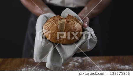 Extending flour-dusted hands holding round artisan loaf on gray cloth in bakery wearing apron 135575866