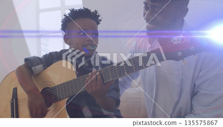 Smiling father watching son playing acoustic guitar on sofa in living room, with lens flare Smiling father watching son playing acoustic guitar on sofa in living room, with lens flare 135575867