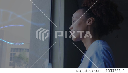Gazing African American nurse in blue scrubs out corridor window, viewing facade, trees, copy space 135576341