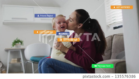 Holding mother leaning forward kissing baby cheek on couch at home, with notification overlays 135576386