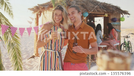 Smiling couple holding cocktail with lime wedge and beer bottle at tiki bar, with colorful bunting Smiling couple holding cocktail with lime wedge and beer bottle at tiki bar, with colorful bunting 135576396