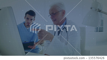 Physician in white coat and trainee in scrubs reviewing monitor in clinic office, with clipboard Physician in white coat and trainee in scrubs reviewing monitor in clinic office, with clipboard 135576426
