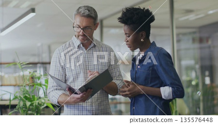 Reviewing black binder man in checkered shirt and woman in denim in modern office, with eyeglasses Reviewing black binder man in checkered shirt and woman in denim in modern office, with eyeglasses 135576448