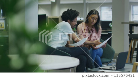 Two coworkers reviewing printed documents in office lounge, with slim laptop and glass of water Two coworkers reviewing printed documents in office lounge, with slim laptop and glass of water 135576451