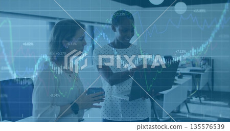Analyzing charts, two businesswomen discussing data in office, with monitors, laptop and coffee mug 135576539