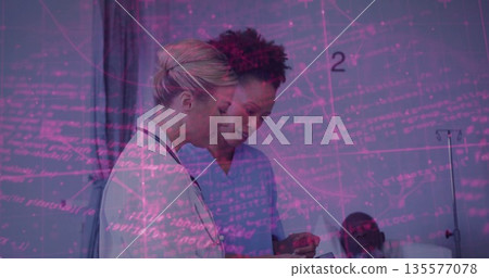 Doctor in white coat and nurse in blue scrubs reviewing tablet in hospital room, with bed 135577078
