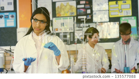 Holding pipette and dish, teenage girl wearing lab coat conducting experiment in science lab 135577088