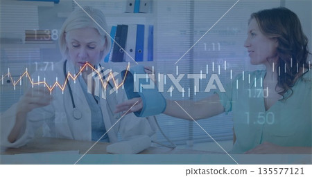 Senior female doctor checking patient blood pressure with cuff on clinic desk, stethoscope, binders Senior female doctor checking patient blood pressure with cuff on clinic desk, stethoscope, binders 135577121