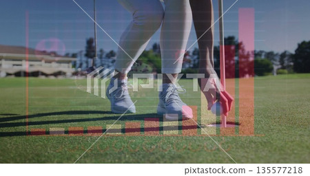 Bending golfer in athletic pants placing golf ball into hole on putting green, with data overlays 135577218