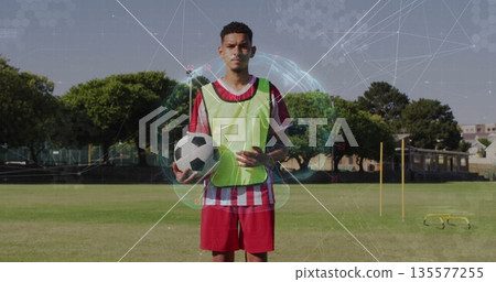 Standing soccer player holding ball wearing neon pinnie near agility poles on training field Standing soccer player holding ball wearing neon pinnie near agility poles on training field 135577255