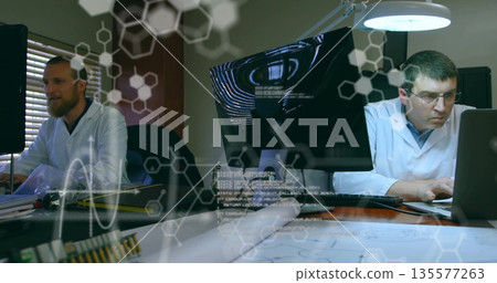 Monitoring two researchers in lab coats typing on laptop and reviewing schematics in lab, with PCBs Monitoring two researchers in lab coats typing on laptop and reviewing schematics in lab, with PCBs 135577263