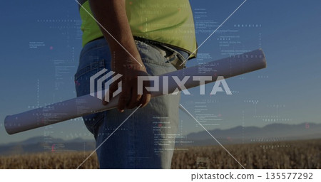 Surveying engineer in high-visibility shirt holding blueprint tube in harvested field, data overlay Surveying engineer in high-visibility shirt holding blueprint tube in harvested field, data overlay 135577292