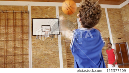 Shooting teenage boy in blue jersey taking shot toward hoop at school gym, with second basketball Shooting teenage boy in blue jersey taking shot toward hoop at school gym, with second basketball 135577309