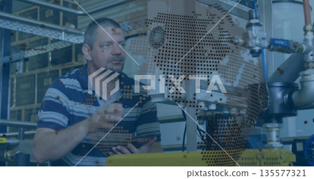Standing male technician gesturing towards machinery in factory, with tablet and world map overlay Standing male technician gesturing towards machinery in factory, with tablet and world map overlay 135577321