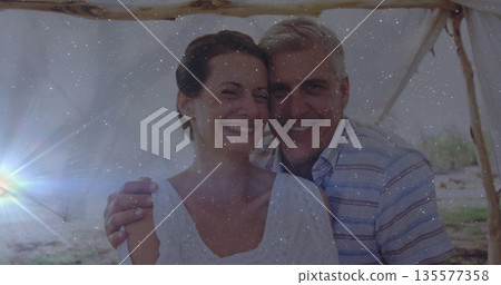 Smiling mid adult couple standing under white cloth canopy by lakeside, with wooden poles Smiling mid adult couple standing under white cloth canopy by lakeside, with wooden poles 135577358