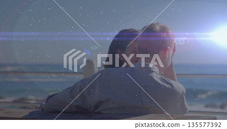 Sitting couple leaning into each other on wood railing overlooking ocean, sun lens flare 135577392