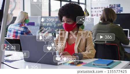 Collaborating masked colleagues sharing open office using laptops, with floating notification icons Collaborating masked colleagues sharing open office using laptops, with floating notification icons 135577425
